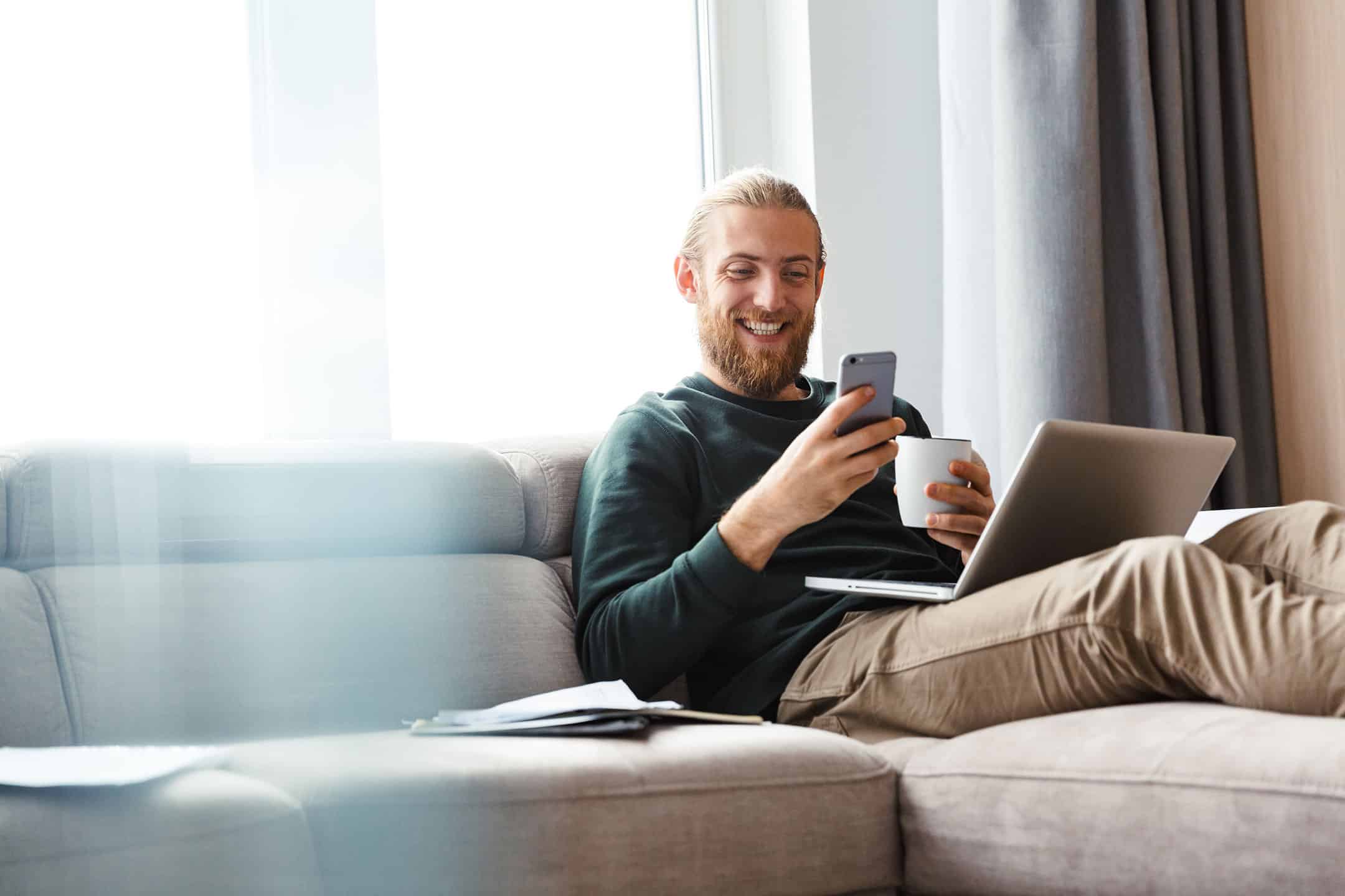 happy man sitting on couch with laptop and phone in hand