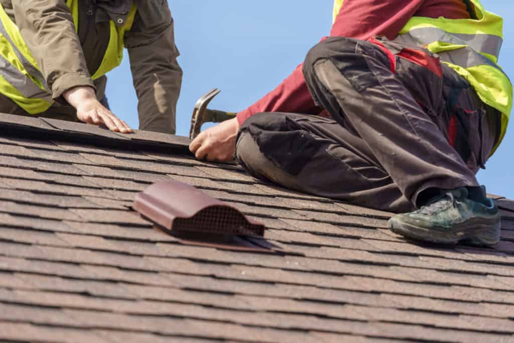 Two mature roofers in protective uniform using helmet and installing asphalt shingle or roof tile on top of new house against blue sky