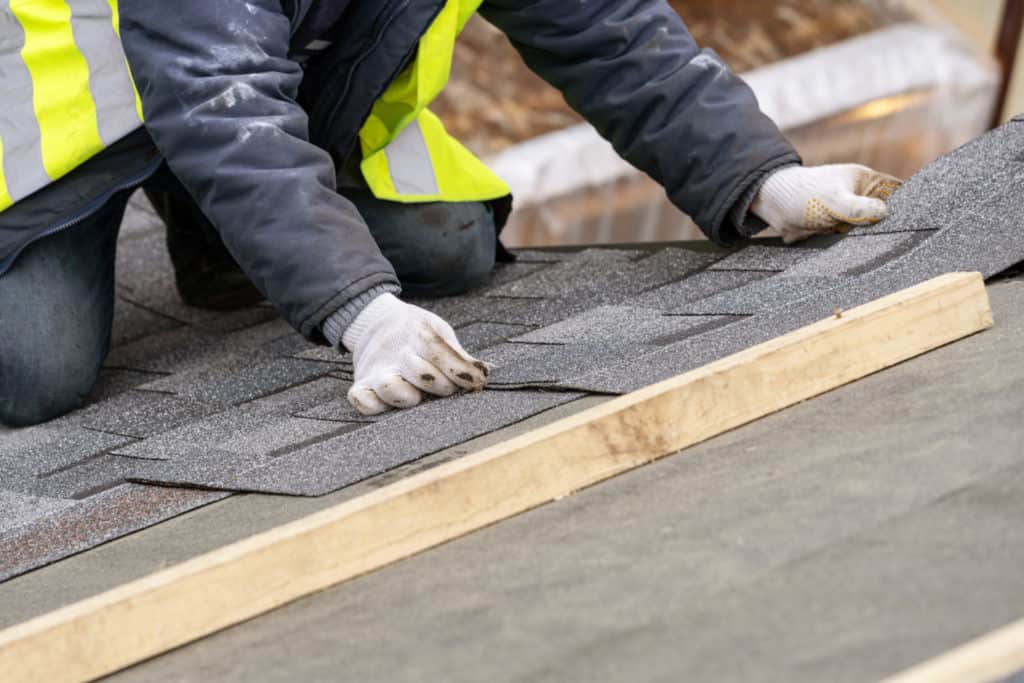Mature and skilled workman in uniform work wear installing asphalt or bitumen tile on top of the roof under construction house
